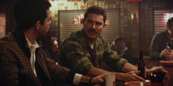 Movie still from “The Greatest Beer Run Ever” (2022), directed by Peter Farrelly – A man sitting at a table with another man in the background; Medium shot, Over the shoulder angle