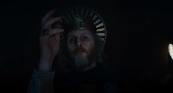 Movie still from “The Green Knight” (2021), directed by David Lowery – A man with a beard wearing a crown and holding a cigarette; Close Up shot, Low angle