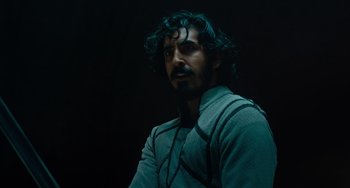 Movie still from “The Green Knight” (2021), directed by David Lowery – A man with long curly hair is looking at the camera; Close Up shot, Over the shoulder angle
