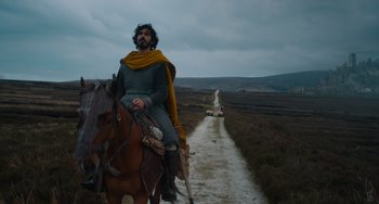 Movie still from “The Green Knight” (2021), directed by David Lowery – A man riding a horse down a dirt road; Wide shot, Low angle