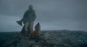 Movie still from “The Green Knight” (2021), directed by David Lowery – A man standing next to a fox on a rocky hill; Wide shot, Low angle