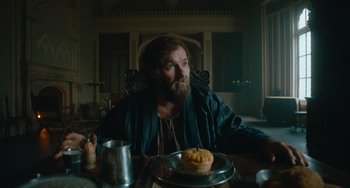 Movie still from “The Green Knight” (2021), directed by David Lowery – A man sitting at a table with a pie in front of him; Medium shot, Over the shoulder angle
