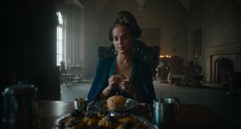 Movie still from “The Green Knight” (2021), directed by David Lowery – A woman sitting in front of a plate of food in a room; Medium shot, Low angle