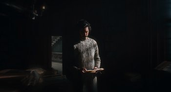 Movie still from “The Green Knight” (2021), directed by David Lowery – A man standing in a dark room holding a book; Medium shot, High angle