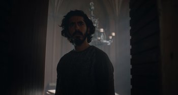 Movie still from “The Green Knight” (2021), directed by David Lowery – A man standing in a dark room with a chandelier in the background; Medium shot, Low angle