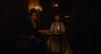 Movie still from “The Green Knight” (2021), directed by David Lowery – Two women sitting at a table in a dimly - lit room; Medium shot, Over the shoulder angle