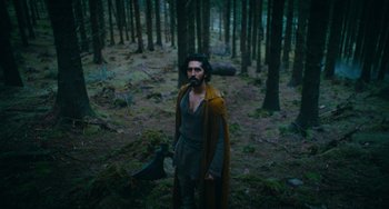 Movie still from “The Green Knight” (2021), directed by David Lowery – A man with a beard and a long coat standing in the woods; Wide shot, Low angle