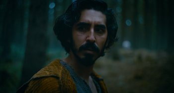 Movie still from “The Green Knight” (2021), directed by David Lowery – A man with long hair and a beard is standing in the woods; Close Up shot, Over the shoulder angle