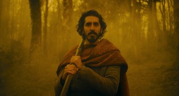 Movie still from “The Green Knight” (2021), directed by David Lowery – A man with a beard holding a stick in the woods; Medium shot, Low angle
