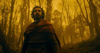 Movie still from “The Green Knight” (2021), directed by David Lowery – A man with a beard is standing in the woods; Medium shot, Low angle