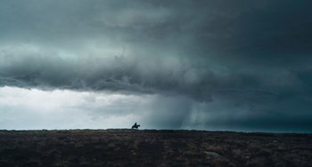 Movie still from “The Green Knight” (2021), directed by David Lowery – A person riding a horse on a field under a cloudy sky; Extreme Wide shot, Low angle