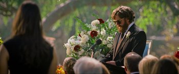 Movie still from “The Hangover Part III” (2013), directed by Todd Phillips – A man in a suit is speaking into a microphone at an event; Medium shot, Over the shoulder angle