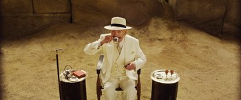 Movie still from “Death on the Nile” (2022), directed by Kenneth Branagh – An older man in a white suit and hat drinking a cup of coffee; Wide shot, High angle