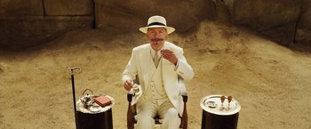 Movie still from “Death on the Nile” (2022), directed by Kenneth Branagh – A man in a white suit and hat sitting in a chair; Medium shot, Low angle