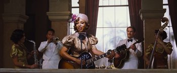 Movie still from “Death on the Nile” (2022), directed by Kenneth Branagh – A woman in a gold dress holding a guitar; Medium shot, Over the shoulder angle