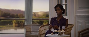 Movie still from “Death on the Nile” (2022), directed by Kenneth Branagh – A woman sitting at a table with a plate of food; Medium shot, Over the shoulder angle