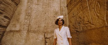 Movie still from “Death on the Nile” (2022), directed by Kenneth Branagh – A woman in a white suit standing in front of an egyptian wall; Medium shot, Low angle
