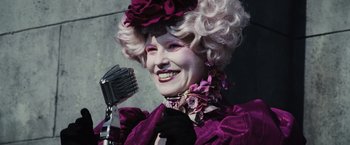 Movie still from “The Hunger Games” (2012), directed by Gary Ross – An older woman in a purple dress with a microphone in front of it; Medium shot, Over the shoulder angle