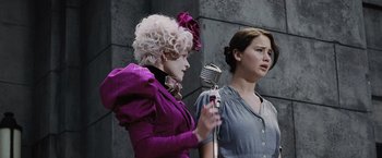 Movie still from “The Hunger Games” (2012), directed by Gary Ross – Two women sing into a microphone in front of a wall; Medium shot, Low angle