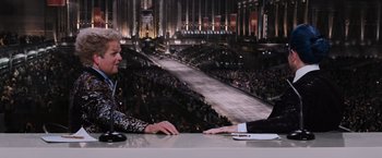 Movie still from “The Hunger Games” (2012), directed by Gary Ross – A man sitting at a table in front of an audience; Medium shot, Over the shoulder angle