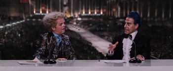 Movie still from “The Hunger Games” (2012), directed by Gary Ross – Two people sitting at a table in front of an audience; Medium shot, Over the shoulder angle