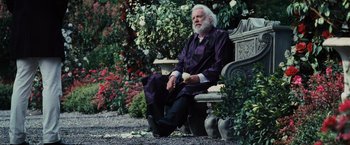 Movie still from “The Hunger Games” (2012), directed by Gary Ross – An older man sitting on a bench in a flower garden; Medium shot, Over the shoulder angle