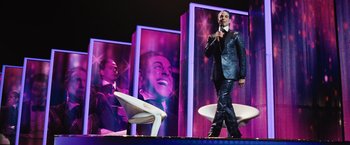 Movie still from “The Hunger Games” (2012), directed by Gary Ross – A man standing on a stage holding a microphone; Wide shot, Low angle