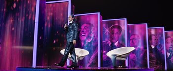 Movie still from “The Hunger Games” (2012), directed by Gary Ross – A man walking on a stage in front of a stage with pictures on the wall; Wide shot, Over the shoulder angle