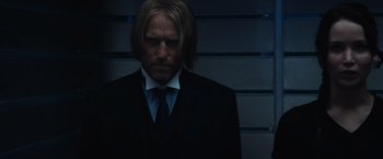 Movie still from “The Hunger Games” (2012), directed by Gary Ross – A man wearing a suit and tie standing in front of a wall; Close Up shot, Over the shoulder angle