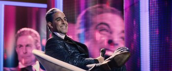 Movie still from “The Hunger Games” (2012), directed by Gary Ross – A man sitting in front of a microphone on a stage; Medium shot, Over the shoulder angle