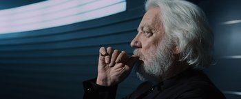 Movie still from “The Hunger Games” (2012), directed by Gary Ross – An older man with white hair and a white beard is holding his hand to his mouth; Close Up shot, Over the shoulder angle