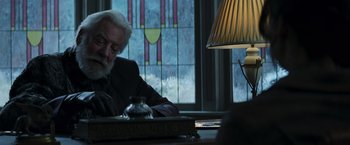 Movie still from “The Hunger Games: Catching Fire” (2013), directed by Francis Lawrence – An older man sitting at a table in front of a stained glass window; Medium shot, Over the shoulder angle