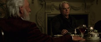 Movie still from “The Hunger Games: Catching Fire” (2013), directed by Francis Lawrence – A man sitting at a table in front of another man holding a plate; Medium shot, Over the shoulder angle
