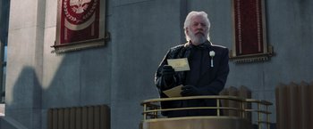 Movie still from “The Hunger Games: Catching Fire” (2013), directed by Francis Lawrence – An older man holding a piece of paper while standing in front of a building; Wide shot, Low angle
