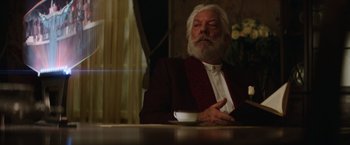 Movie still from “The Hunger Games: Catching Fire” (2013), directed by Francis Lawrence – An older man sitting at a table with a cup of coffee; Medium shot, Low angle