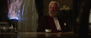 Movie still from “The Hunger Games: Catching Fire” (2013), directed by Francis Lawrence – An older man sitting at a table with a cup of coffee; Medium shot, Over the shoulder angle