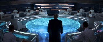 Movie still from “The Hunger Games: Catching Fire” (2013), directed by Francis Lawrence – A man standing in front of a group of people in a room; Wide shot, Over the shoulder angle