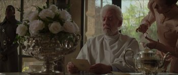 Movie still from “The Hunger Games: Mockingjay - Part 1” (2014), directed by Francis Lawrence – An older man sitting at a table holding a card; Medium shot, Over the shoulder angle