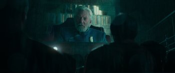 Movie still from “The Hunger Games: Mockingjay - Part 1” (2014), directed by Francis Lawrence – An older man sitting in a chair with a banana in front of him; Medium shot, Over the shoulder angle