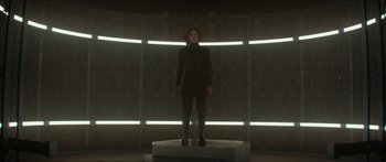 Movie still from “The Hunger Games: Mockingjay - Part 1” (2014), directed by Francis Lawrence – A woman in a black suit stands on a pedestal in front of a wall; Wide shot, Low angle