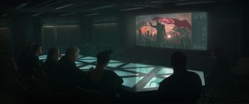 Movie still from “The Hunger Games: Mockingjay - Part 1” (2014), directed by Francis Lawrence – A group of people sitting in front of a large screen; Wide shot, Over the shoulder angle