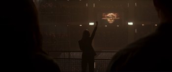 Movie still from “The Hunger Games: Mockingjay - Part 1” (2014), directed by Francis Lawrence – A woman standing in front of an audience holding her arm up in the air; Wide shot, Low angle