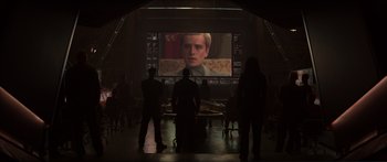Movie still from “The Hunger Games: Mockingjay - Part 1” (2014), directed by Francis Lawrence – A group of people standing in front of a large screen tv; Wide shot, Over the shoulder angle