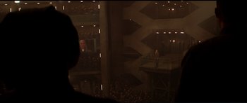 Movie still from “The Hunger Games: Mockingjay - Part 1” (2014), directed by Francis Lawrence – An audience is watching a man stand on a stage; Extreme Wide shot, Over the shoulder angle