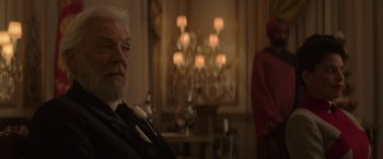 Movie still from “The Hunger Games: Mockingjay - Part 2” (2015), directed by Francis Lawrence – An older man in a suit and tie in front of chandeliers; Medium shot, Over the shoulder angle
