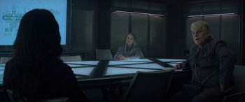 Movie still from “The Hunger Games: Mockingjay - Part 2” (2015), directed by Francis Lawrence – A woman sitting at a table in front of a window; Medium shot, Over the shoulder angle