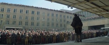 Movie still from “The Hunger Games: Mockingjay - Part 2” (2015), directed by Francis Lawrence – A crowd of people standing in front of a building; Wide shot, Over the shoulder angle