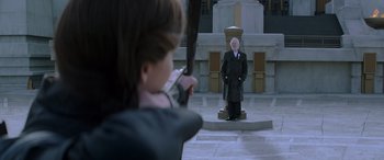 Movie still from “The Hunger Games: Mockingjay - Part 2” (2015), directed by Francis Lawrence – A person taking a picture of a statue of a man in a suit; Wide shot, Over the shoulder angle