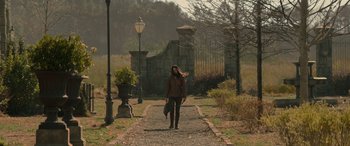 Movie still from “The Hunger Games: Mockingjay - Part 2” (2015), directed by Francis Lawrence – A woman walking down a dirt path holding an umbrella; Wide shot, Over the shoulder angle
