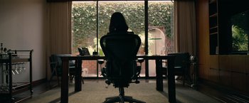 Movie still from “The Hunt” (2020), directed by Craig Zobel – A woman sitting at a desk in front of a large window; Wide shot, Over the shoulder angle